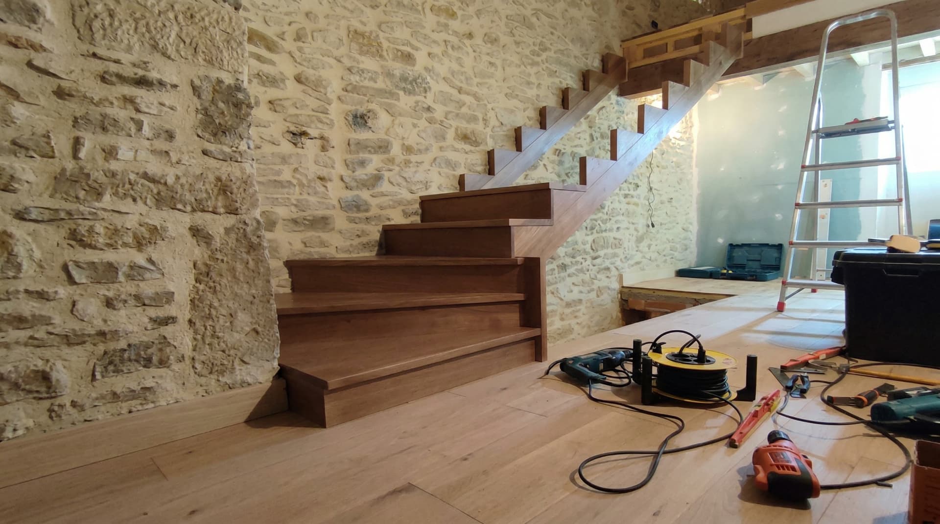 Vista lateral de la instalación de escalera de madera con peldaños en tono oscuro sobre una pared de piedra natural, mostrando herramientas de carpintería y cables en el suelo durante el proceso de montaje profesional.